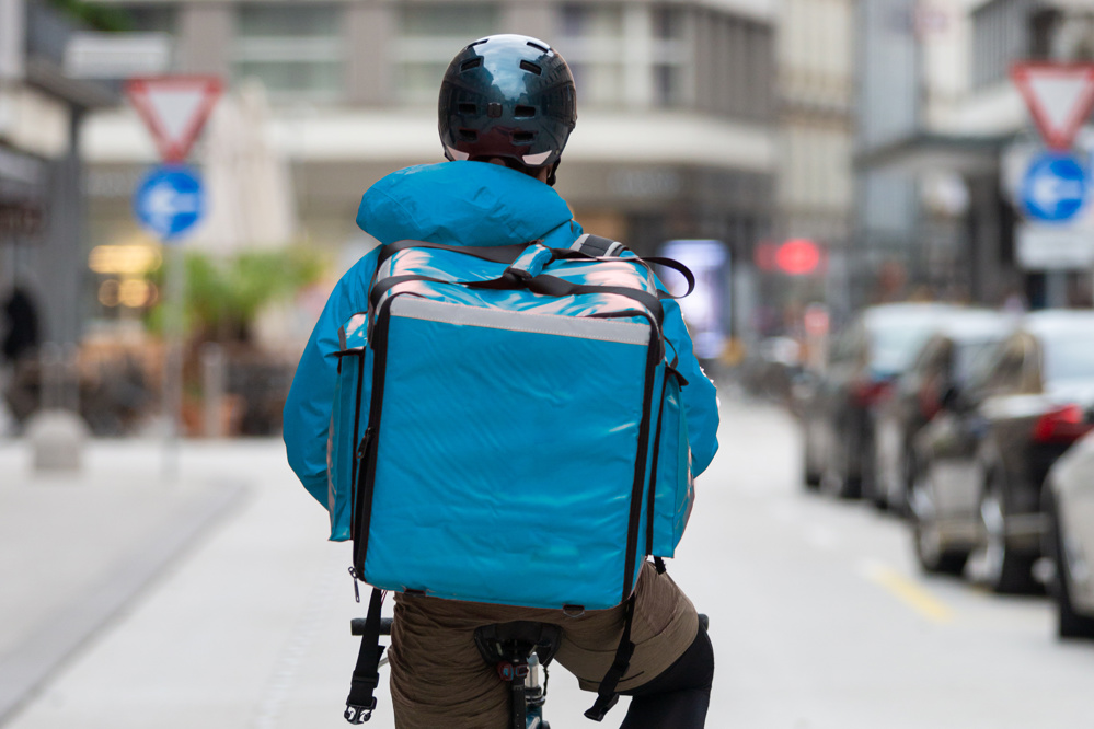 View from behind a delivery rider on a bike, wearing a blue rain jacket and blue food delivery bike, on an urban street.