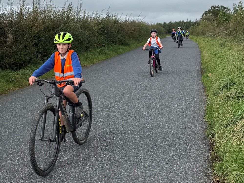 Pupils at Springholm Primary School in Dumfries and Galloway take part in an on-road Bikeability Scotland cycle training session. The pupils can be seen smiling as they ride their bikes towards the camera on a rural, single track road close to their school.