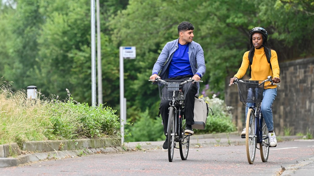 A man and woman talk to each other cycle together on a dedicated cycle path, on a neighbourhood street. Trees and a bus stop can be seen in the background.